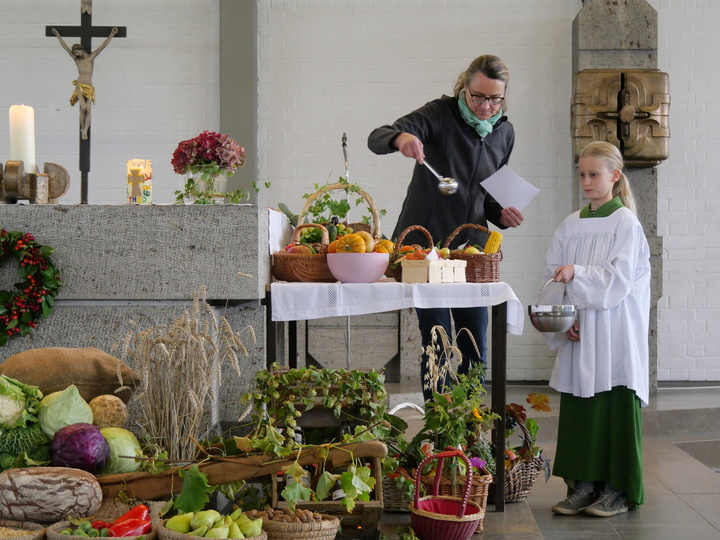 Die Wortgottesdienstleiterin besprengt die Erntegaben mit Weihwasser. Neben ihr steht eine Ministrantin.